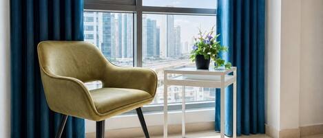 A room with a view of a cityscape through large windows, a single chair, a small table, and a potted plant.