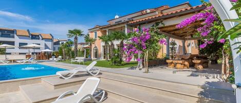 A pool area with lounge chairs, a water feature, and a building with a tiled roof and purple flowers.