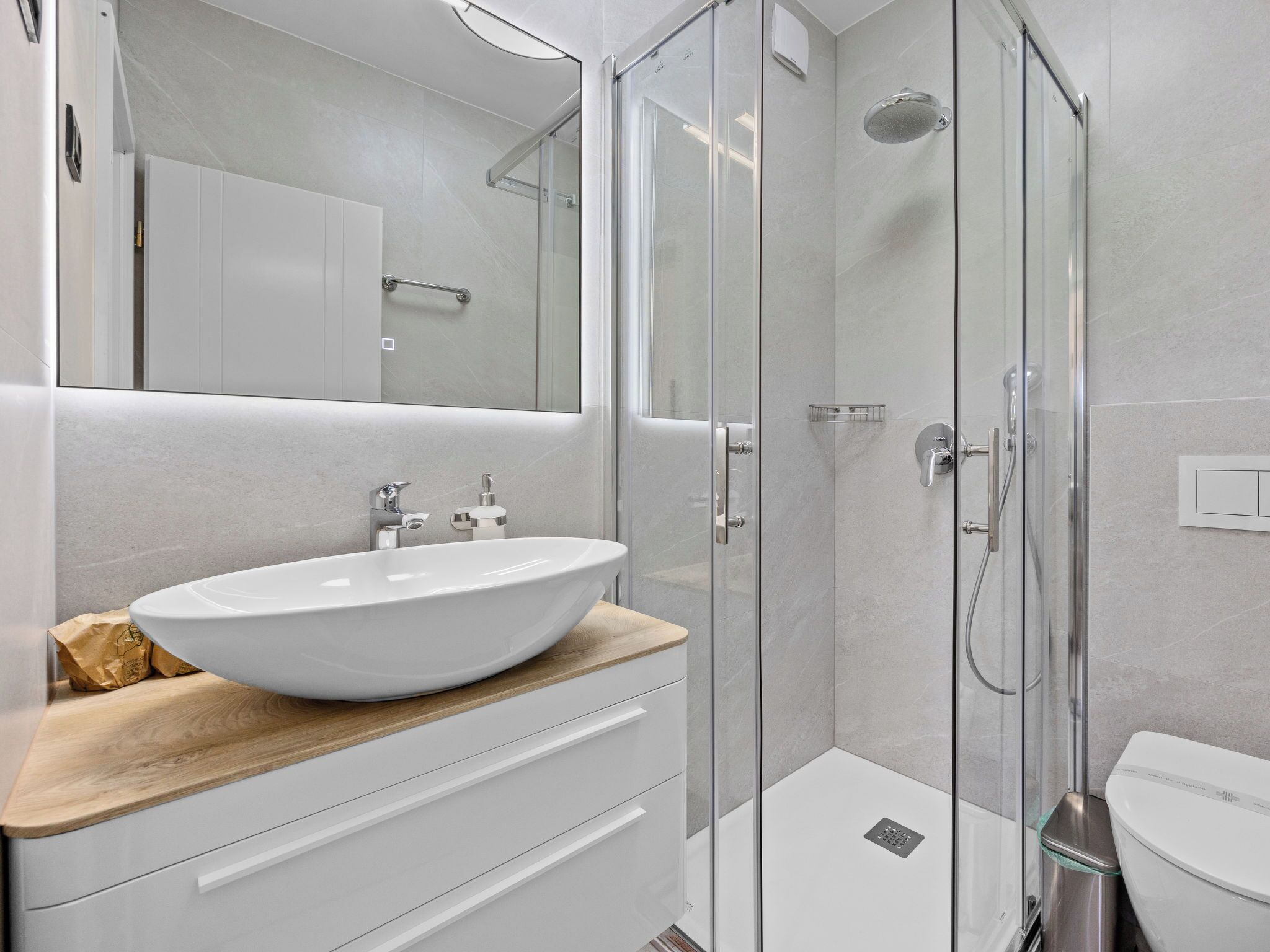 Een moderne badkamer met een groot wit wastafelblad, een glazen douchecabine en een houten wastafelmeubel.