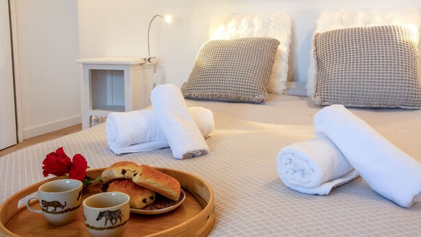 Un lit bien fait, avec des draps blancs, deux serviettes enroulées et un plateau contenant du pain et deux tasses.