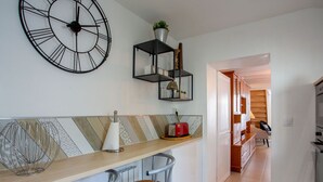 Une cuisine moderne avec un plan de travail en bois, deux tabourets de bar, une horloge et une étagère ornée d’objets décoratifs.