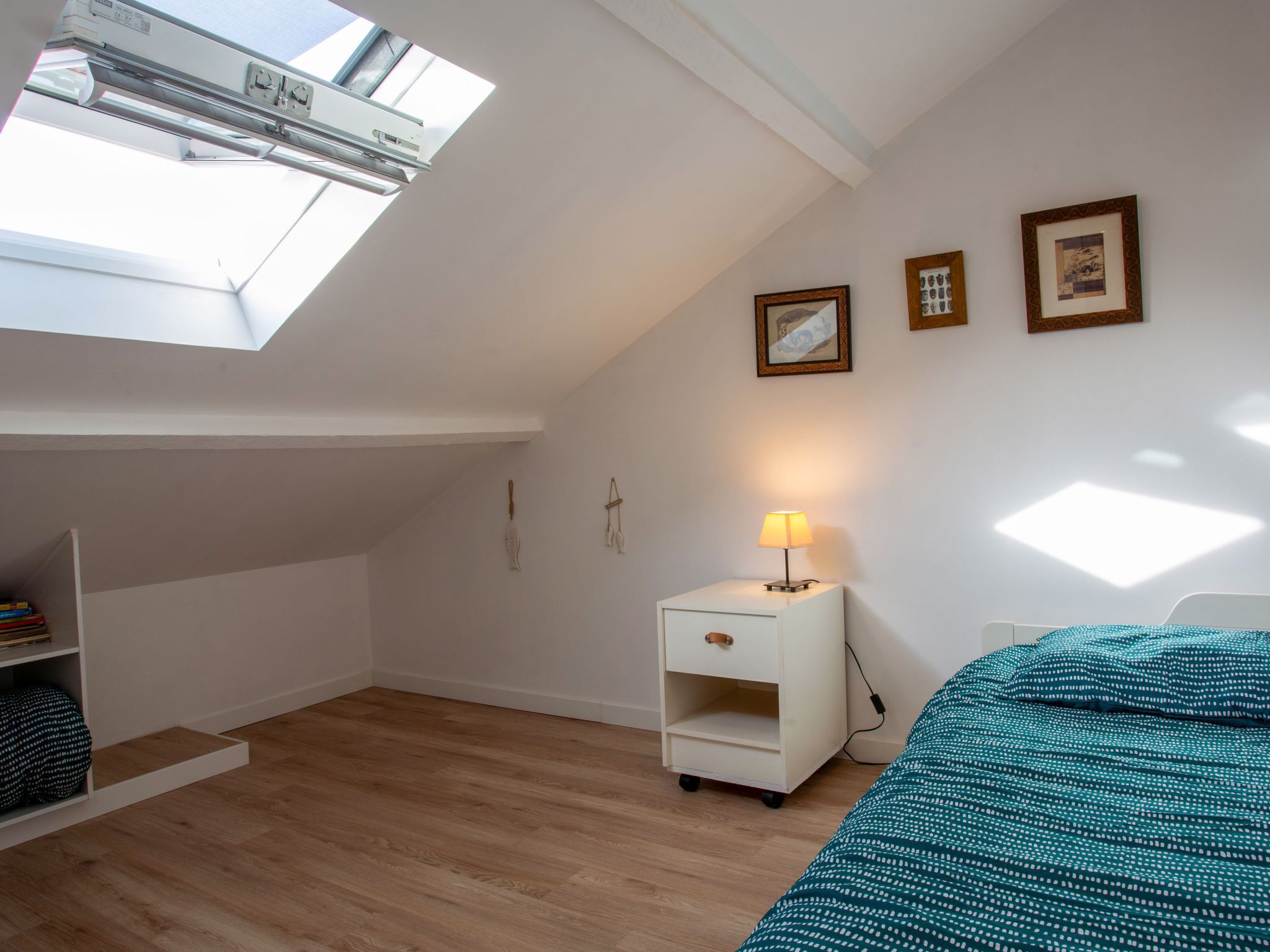 A bedroom with a skylight, a bedside table with a lamp, a bed with a patterned blanket, and a shelf with books.