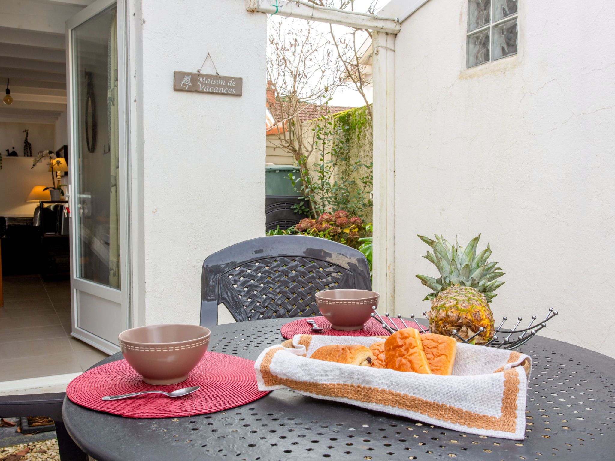 A table set with a bowl, a spoon, a pineapple, and bread on a patio with a chair and a sign reading "Maison de Vacances".