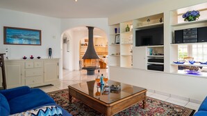 A modern living room with a fireplace, blue sofas, a coffee table, and a kitchen area with shelves and a TV. - Ty Stella by Interhome (Porspoder)