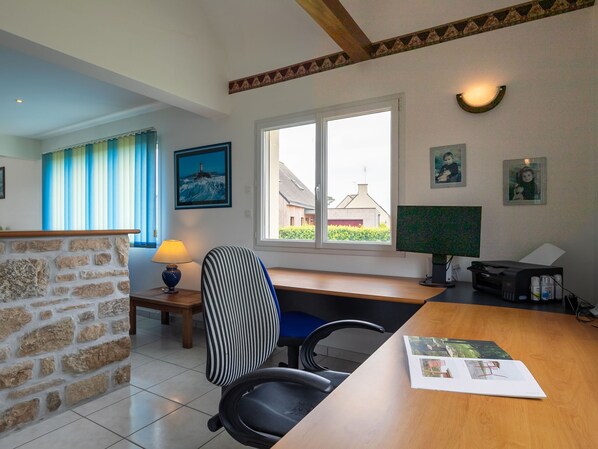 A room with a stone wall, a desk with a computer, a chair, a lamp, and a window with a view of a house.