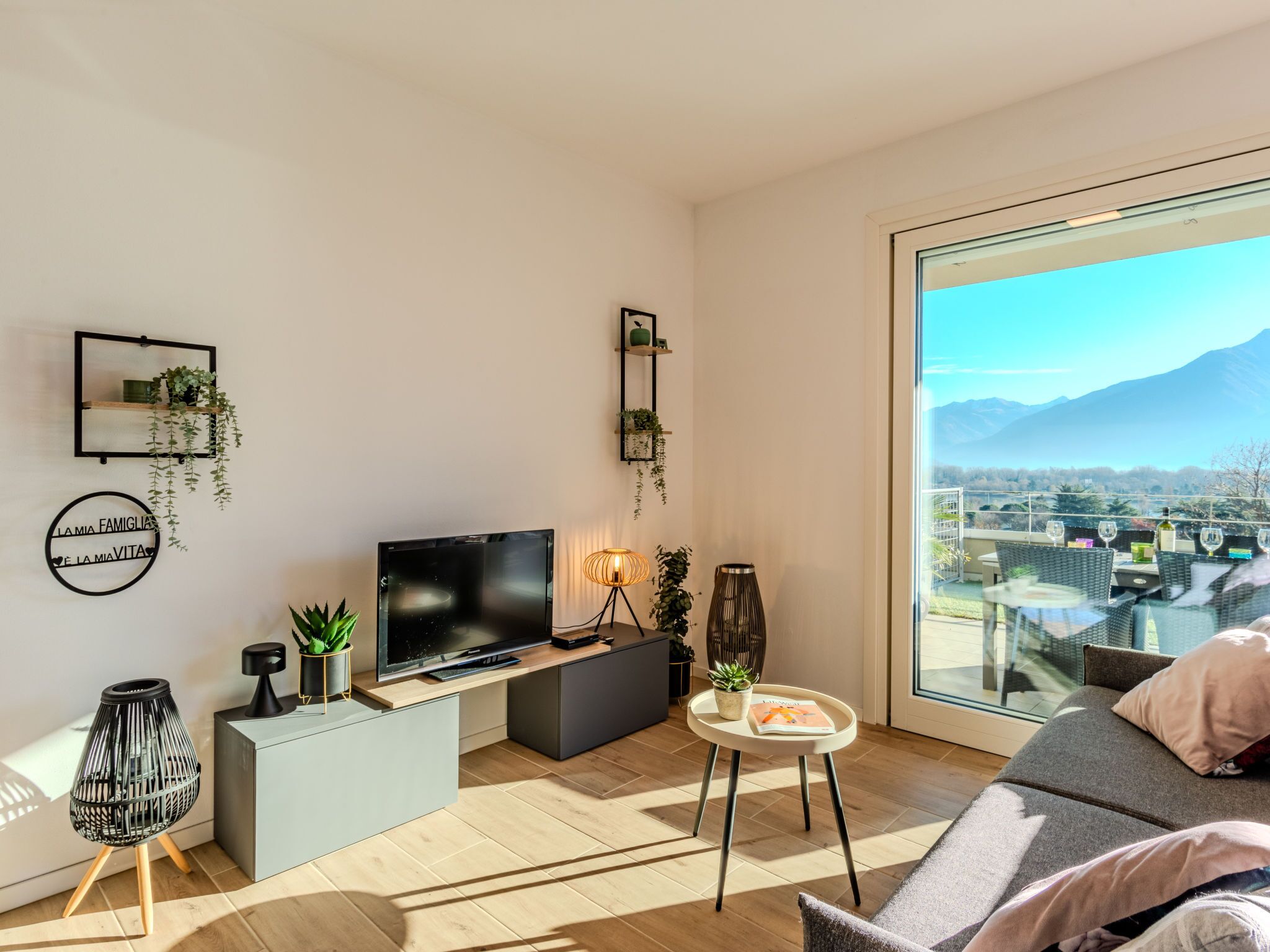 Een moderne woonkamer met een flatscreen-tv, een klein bijzettafeltje en een bank. Er staan planten op planken en een groot raam met uitzicht op de stad.
