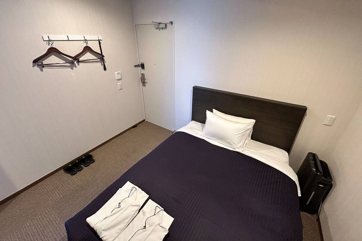 A hotel room with a bed, a coat rack, a door, and a trash bin.
