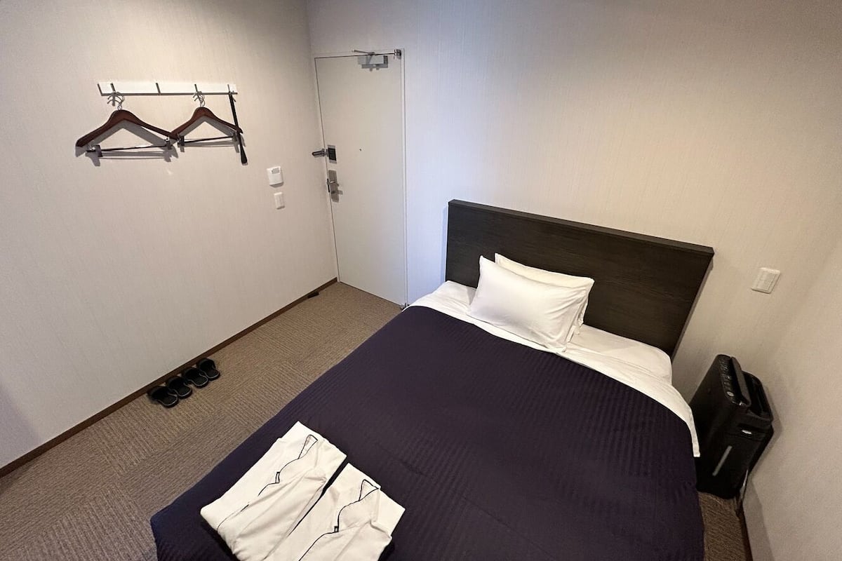 Habitación de hotel con una cama, perchero, puerta y cubo de basura.
