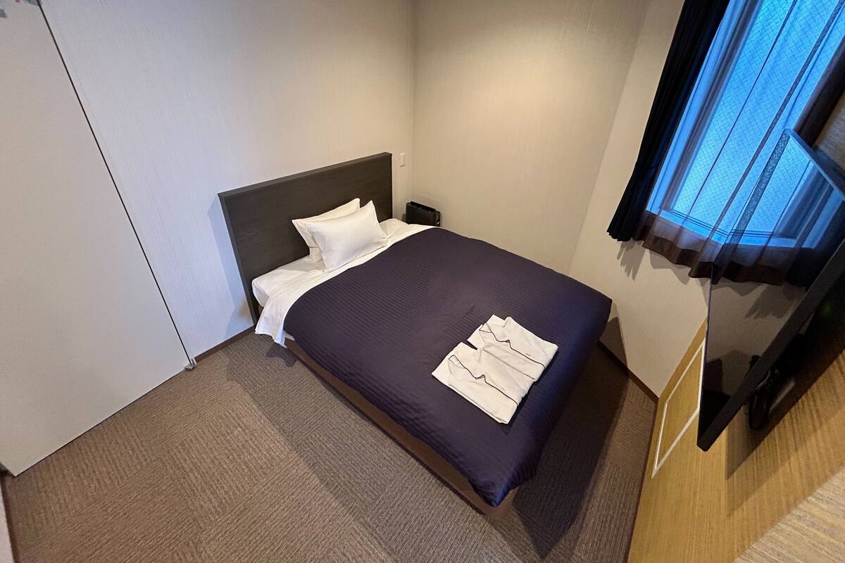 A hotel room with a bed, a flat-screen TV, and a window with curtains.