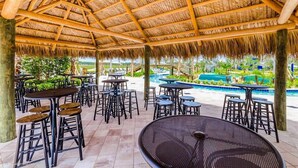 Eine Terrassengaststätte im Freien mit Reetdach, mehreren Tischen und Hockern sowie einem Pool im Hintergrund.
