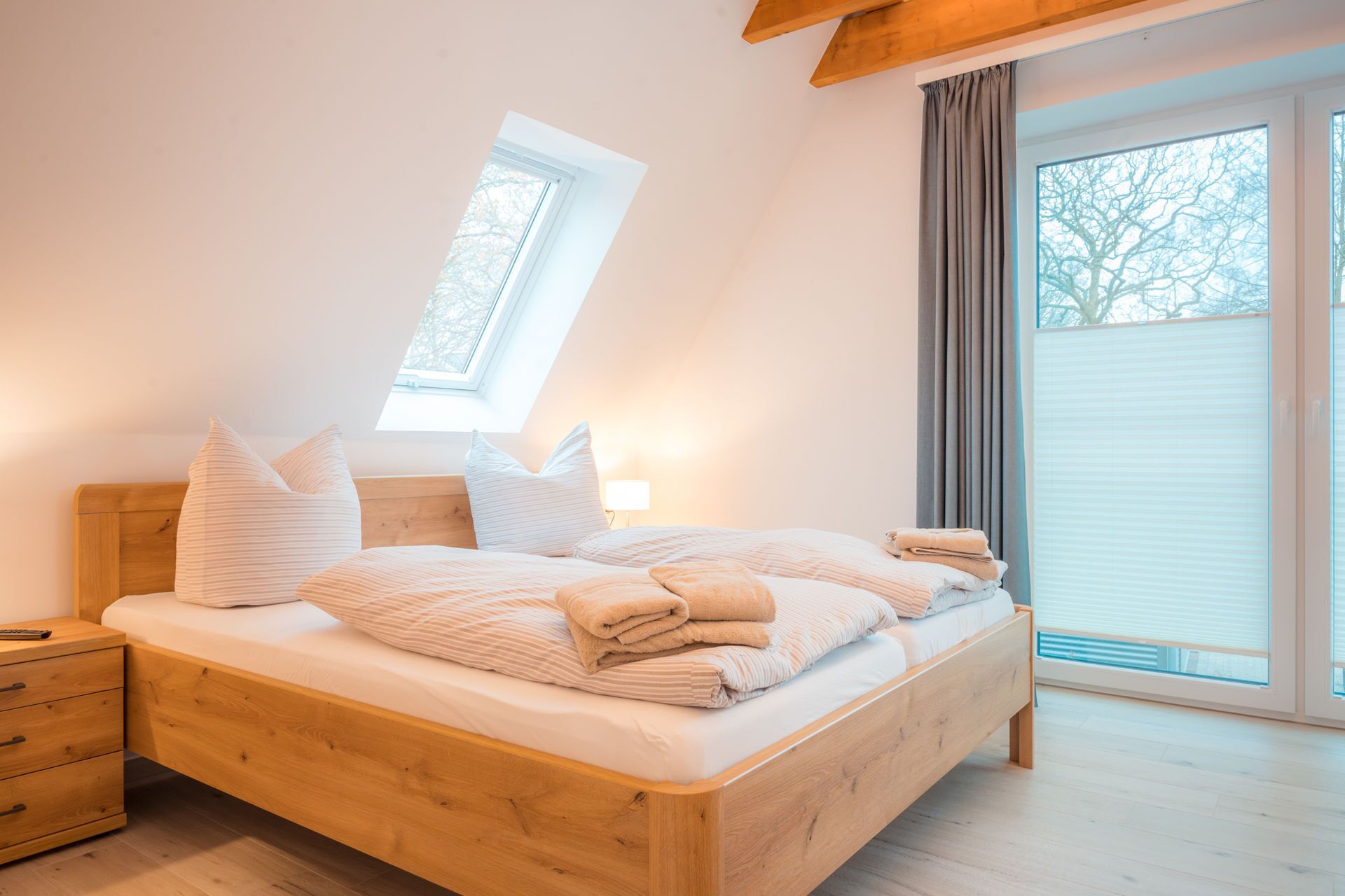 A bedroom with a wooden bed, a nightstand, a bedside lamp, and a window with a view of trees.