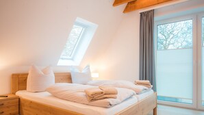 A bedroom with a wooden bed, a nightstand, a bedside lamp, and a window with a view of trees. - "Experience unforgettable days in the Dangaster Düne!" (Dangast)
