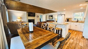 A spacious dining area with a wooden table and chairs, a kitchen with white cabinets, and a large window providing natural light. - 228 Lakeland (Anderson)