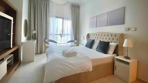 Una habitación de hotel moderna con una cama grande, un televisor, una mesita de noche con lámpara y un ventanal con cortinas.