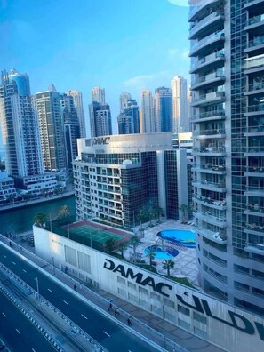 A high-rise building with a prominent DAMAC sign, surrounded by other tall buildings, a road with vehicles, and a swimming pool area.
