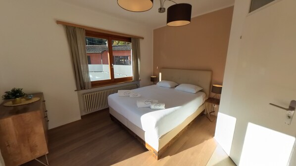 Une chambre à coucher avec un lit, une fenêtre avec des rideaux, un radiateur, une table de chevet avec une lampe et un plancher en bois.
