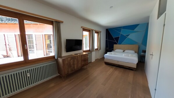 A bedroom with a bed, a TV, a wooden dresser, and large windows.