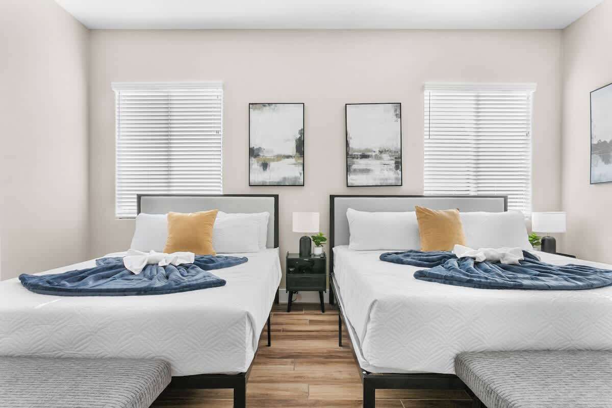 A hotel room with two beds, white bedding, a small bedside table with a lamp, and artwork on the walls.