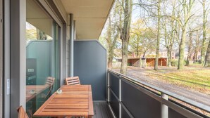 A balcony with a wooden table and chairs, a glass door, and a view of a street and trees. - Vacation apartment "Theos Garten" in Husum for up to 4 people (Husum)