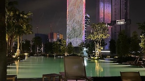 A nighttime view of a hotel pool area with lounge chairs and a reflection of a brightly lit building in the background. - AGILE Infinity Suites TRX By AR (Kuala Lumpur)