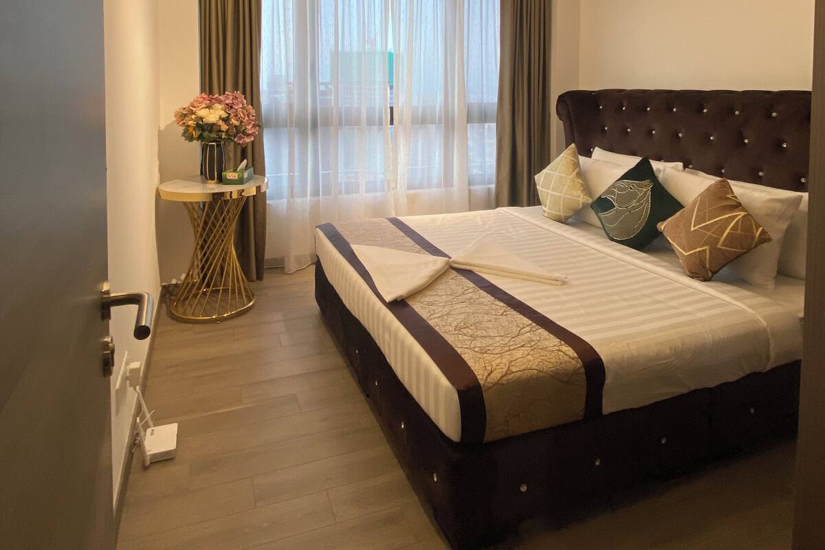 A hotel room with a large bed, a round side table with a vase of flowers, a ceiling light, and a window with curtains.
