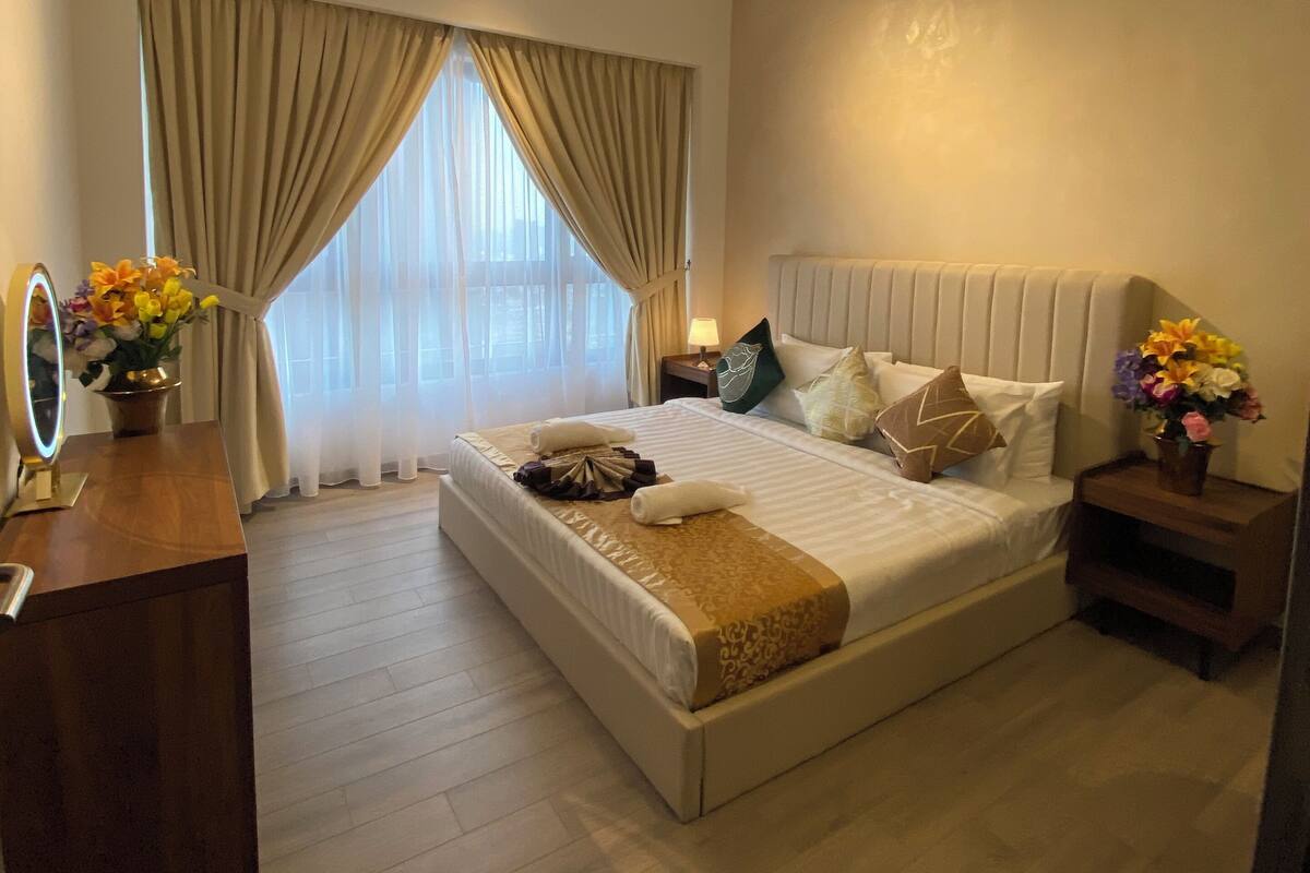 A modern bedroom with a large bed, a ceiling fan, an air conditioning unit, a bedside table with a lamp, and a window with curtains.