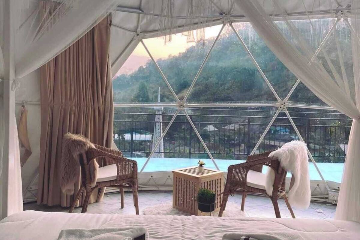 A tented room with a bed, two wicker chairs, a small table, and a view of a pool and trees.