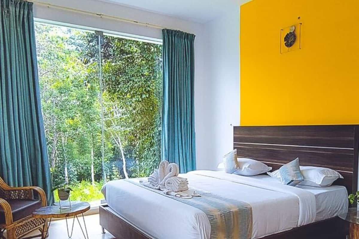 A hotel room with a bed, a chair, a small table, and a view of greenery through the window.