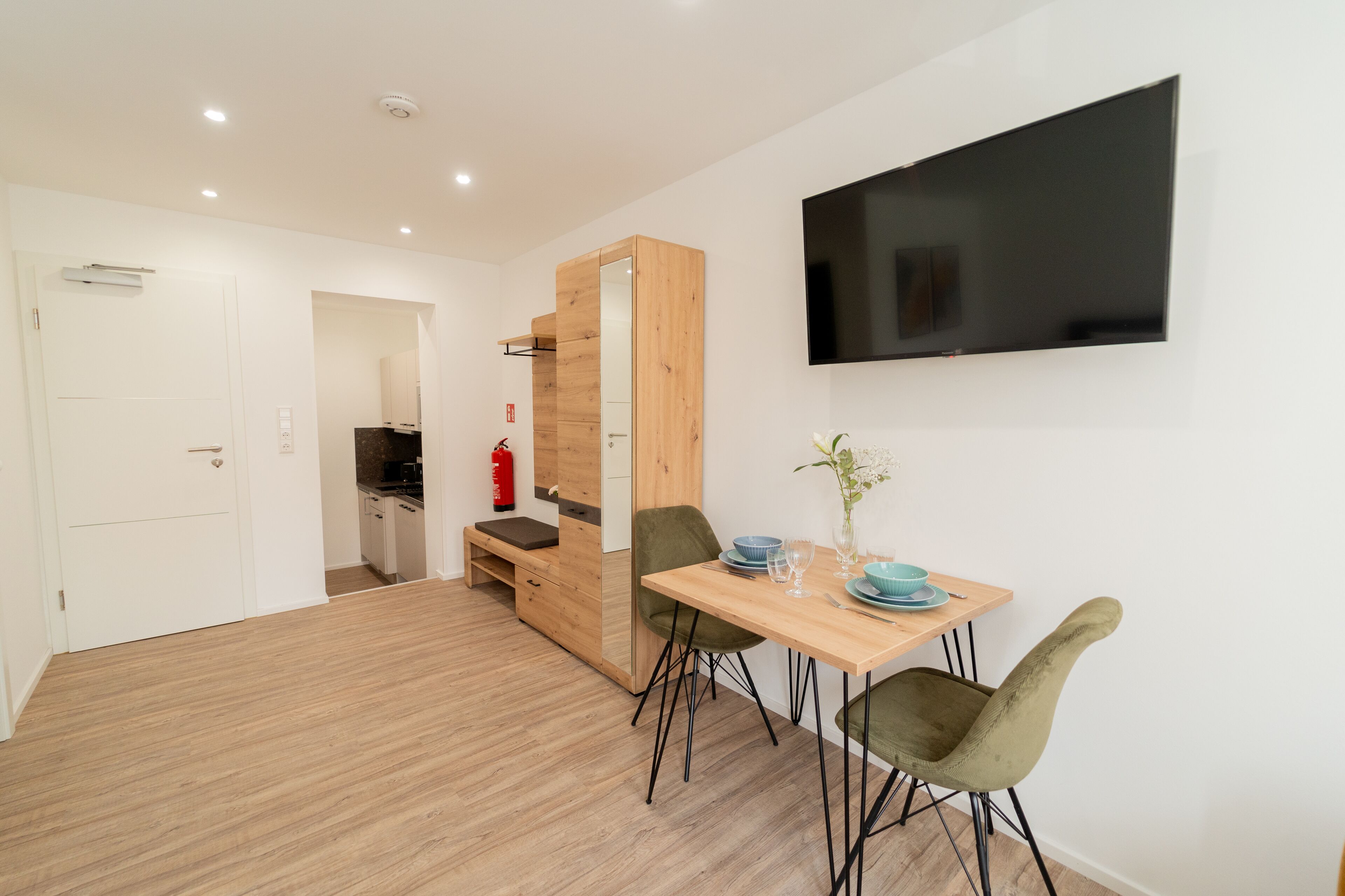A modern kitchen with wooden cabinets, a dining table with chairs, and a flat-screen TV mounted on the wall.