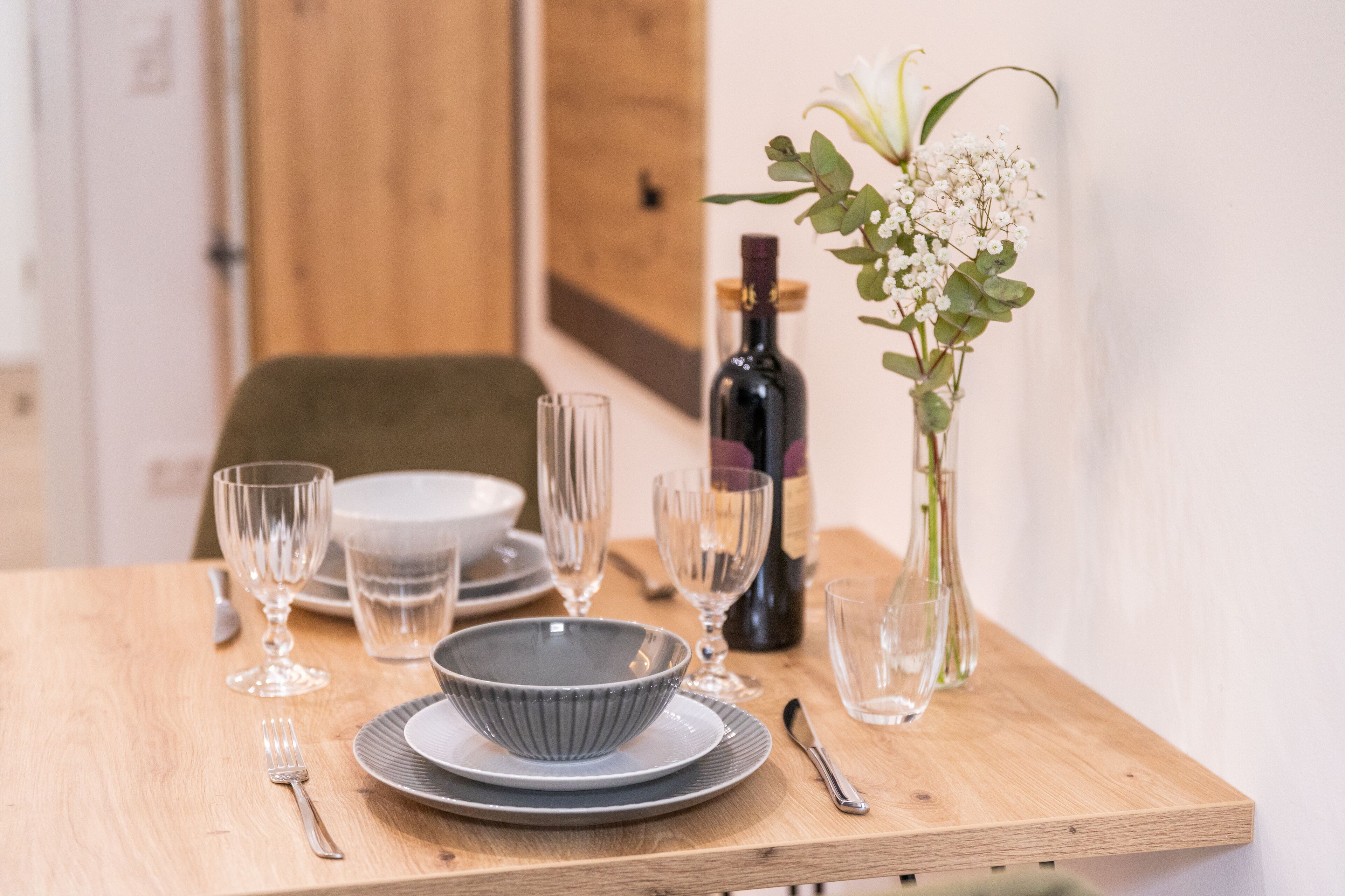 A wooden dining table set with plates, bowls, glasses, cutlery, a bottle, and a vase with flowers.