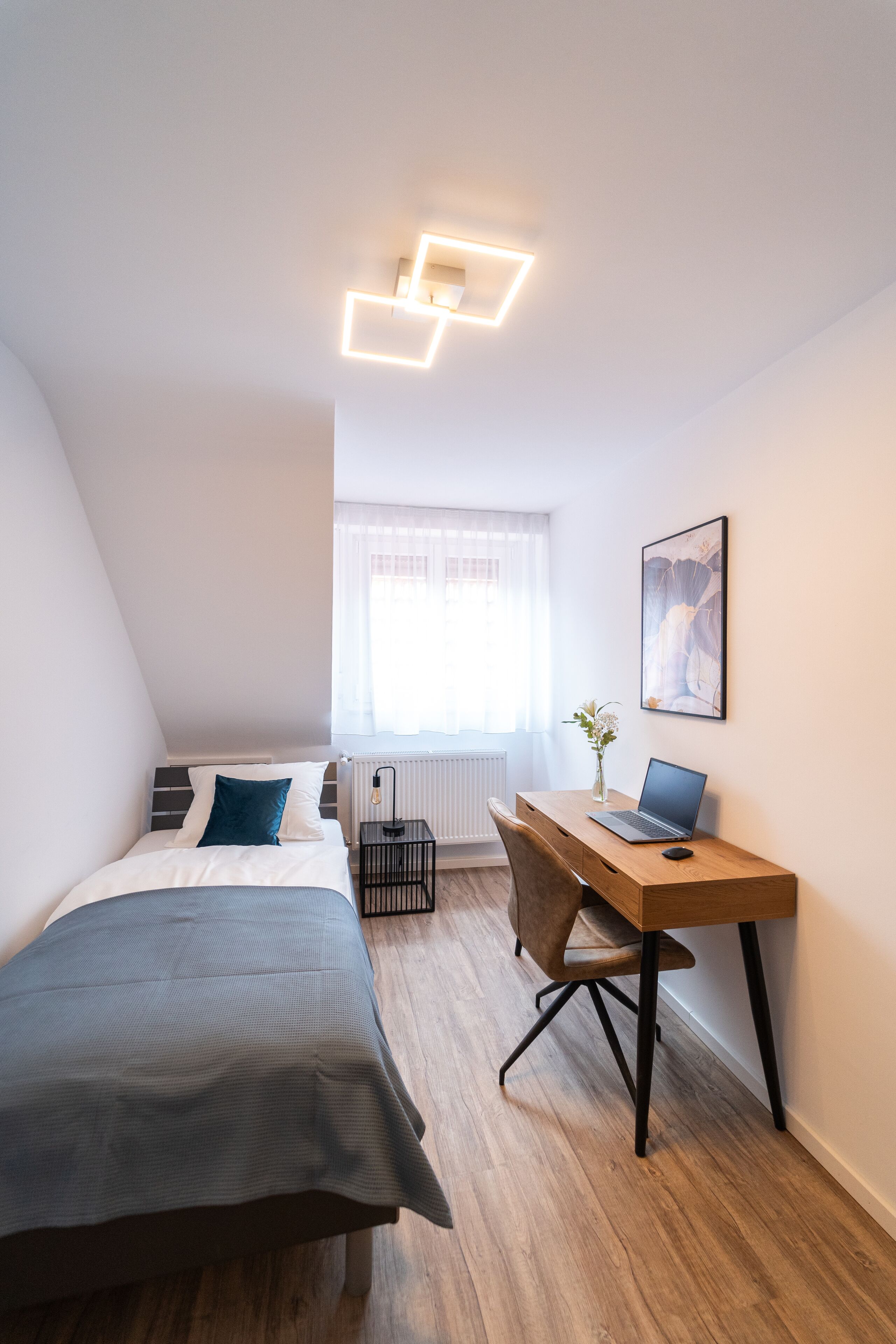 Ein Schlafzimmer mit einem Bett, einem Schreibtisch mit Laptop, einem Stuhl und einem Fenster mit Vorhängen.
