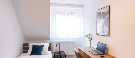 Ein Schlafzimmer mit einem Bett, einem Schreibtisch mit Laptop, einem Stuhl und einem Fenster mit Vorhängen.