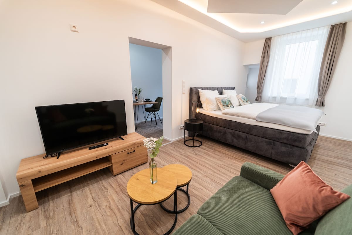 A modern hotel room with a flat-screen TV, a wooden TV stand, a small round table with a vase, a green sofa, and a bed with a grey headboard.