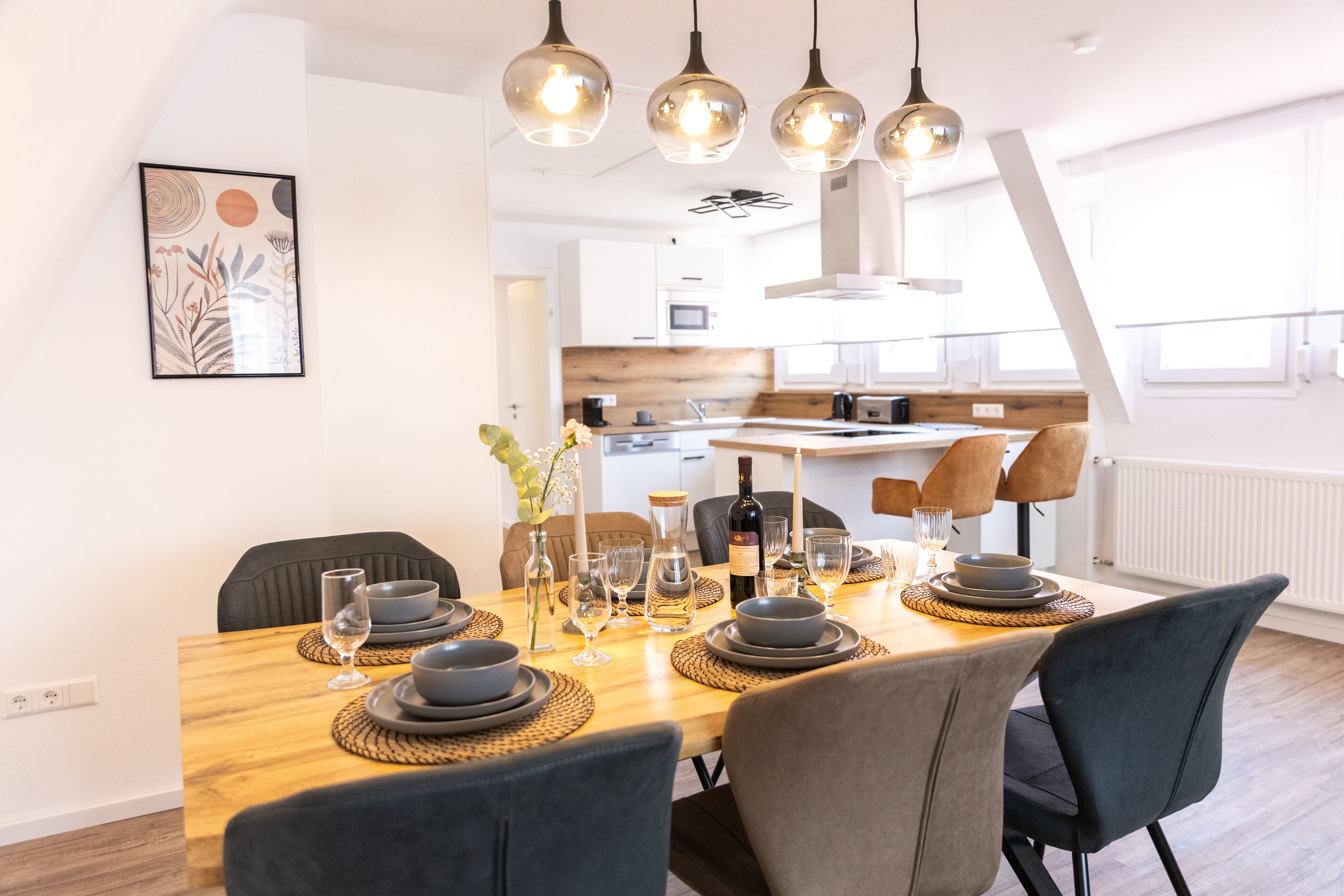 A dining area with a wooden table set for four, surrounded by dark chairs. A kitchen is visible in the background with modern appliances.