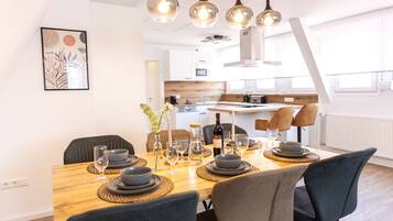 A dining area with a wooden table set for four, surrounded by dark chairs. A kitchen is visible in the background with modern appliances.