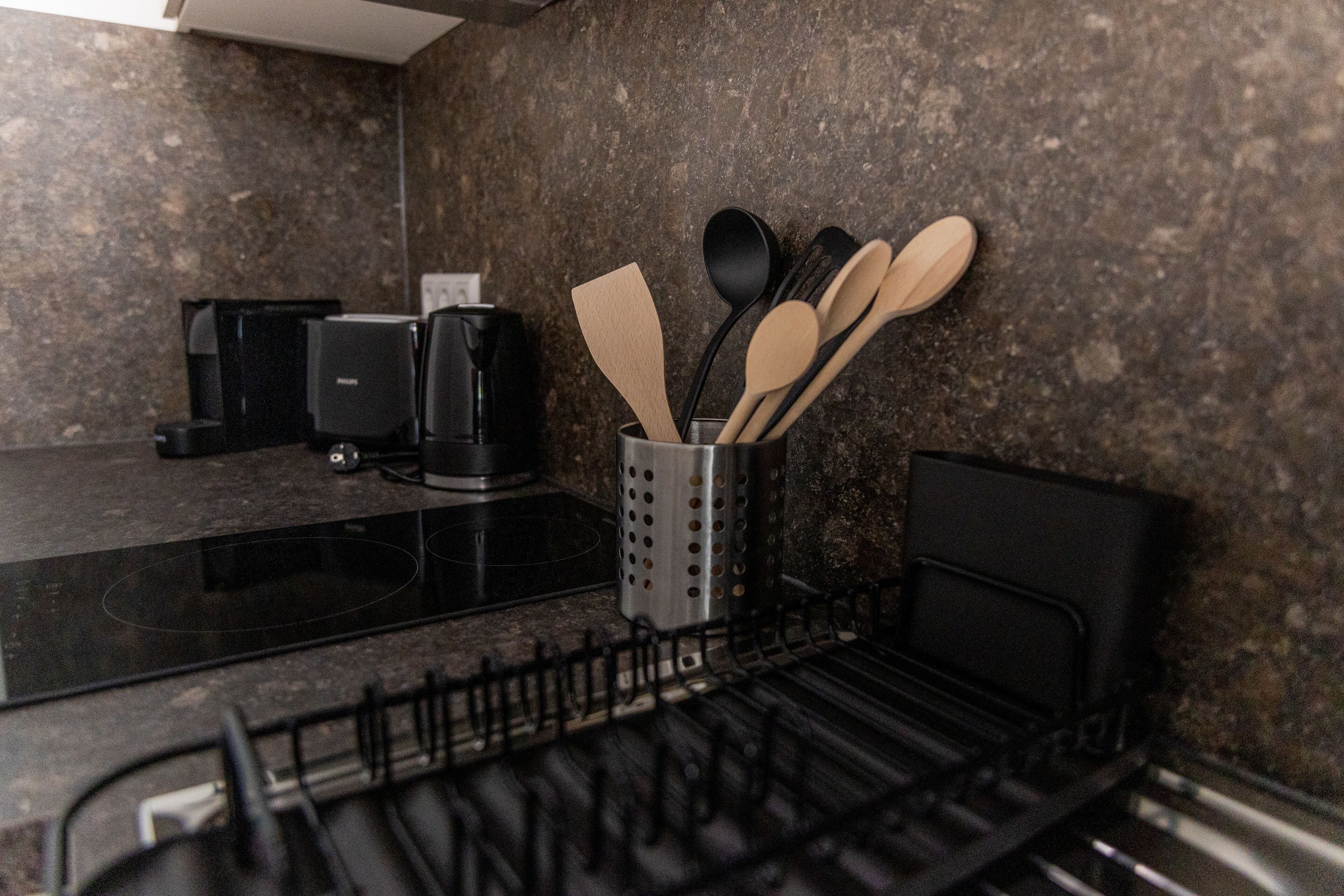 Una cocina con encimera negra, cafetera y utensilios de madera en un organizador.