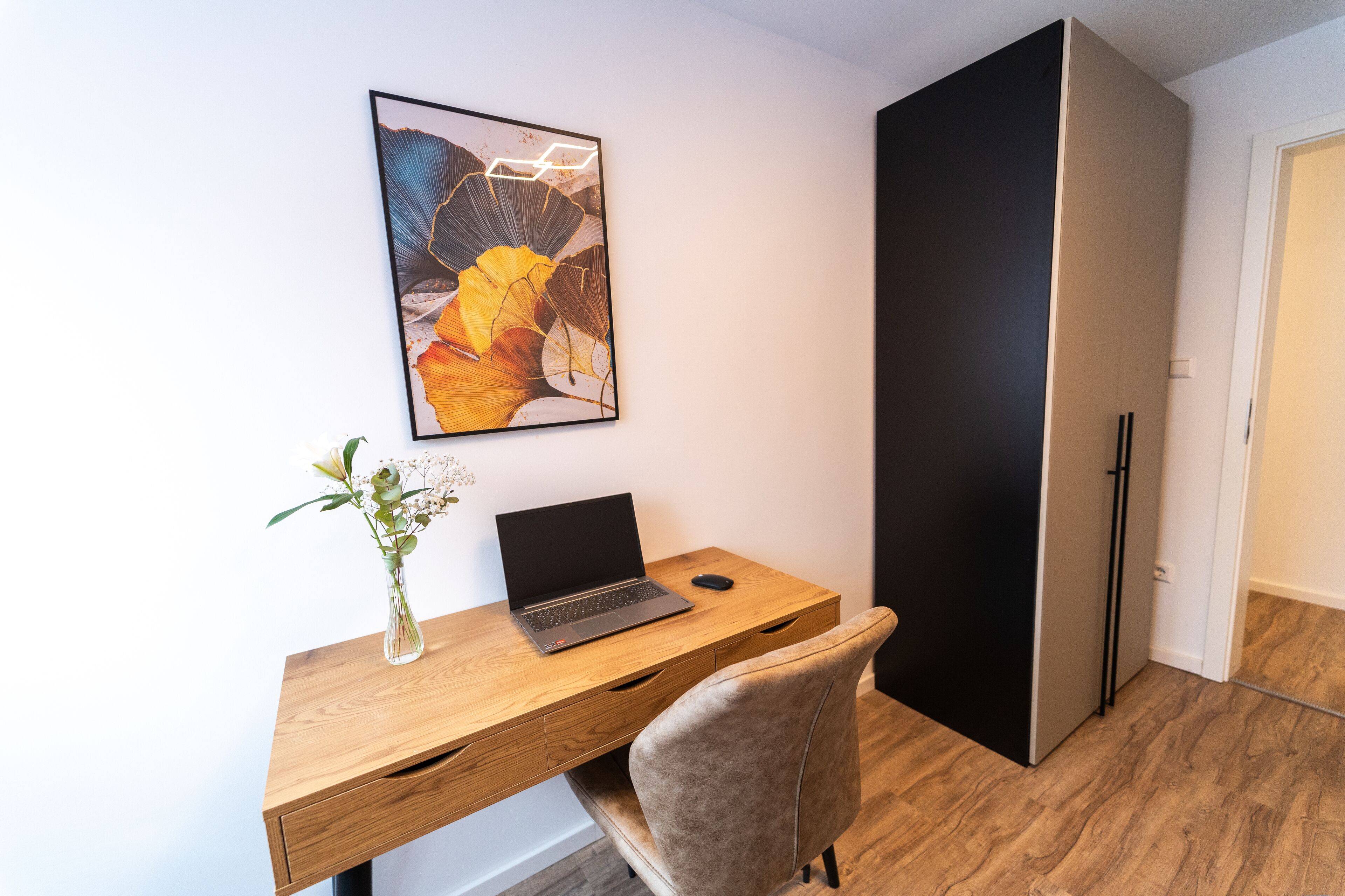 A modern office space with a wooden desk, a laptop, a vase with flowers, and a large abstract painting on the wall.