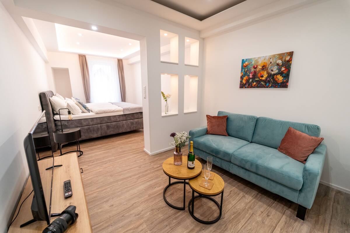 A modern living room with a sofa, a coffee table, a TV, and a bed in the background.