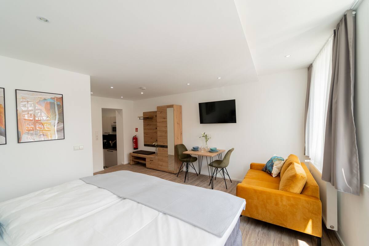 A modern hotel room with a bed, a sofa, a dining table with chairs, a TV, and artwork on the walls.