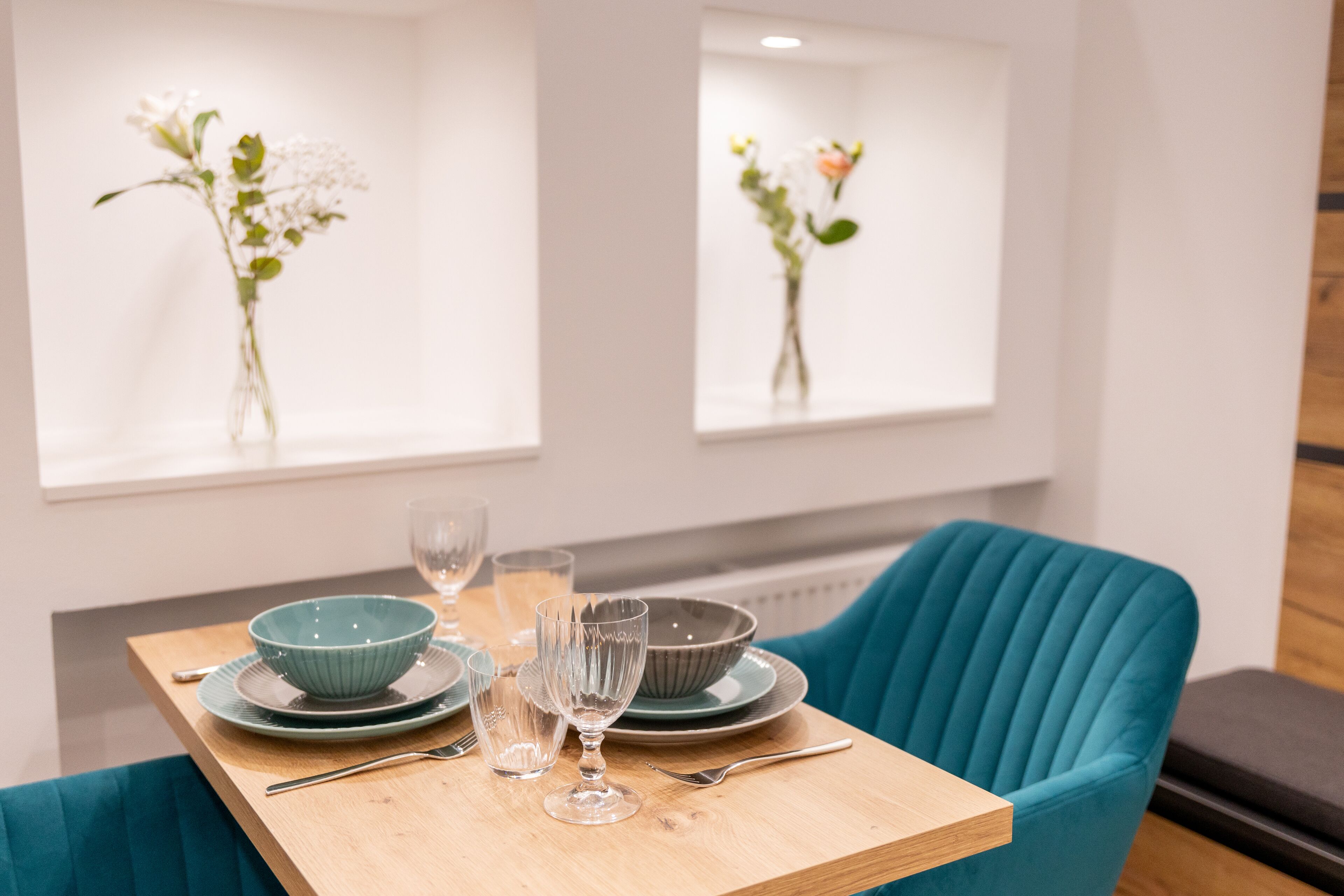 A dining table set with plates, bowls, and glasses, flanked by teal upholstered chairs.