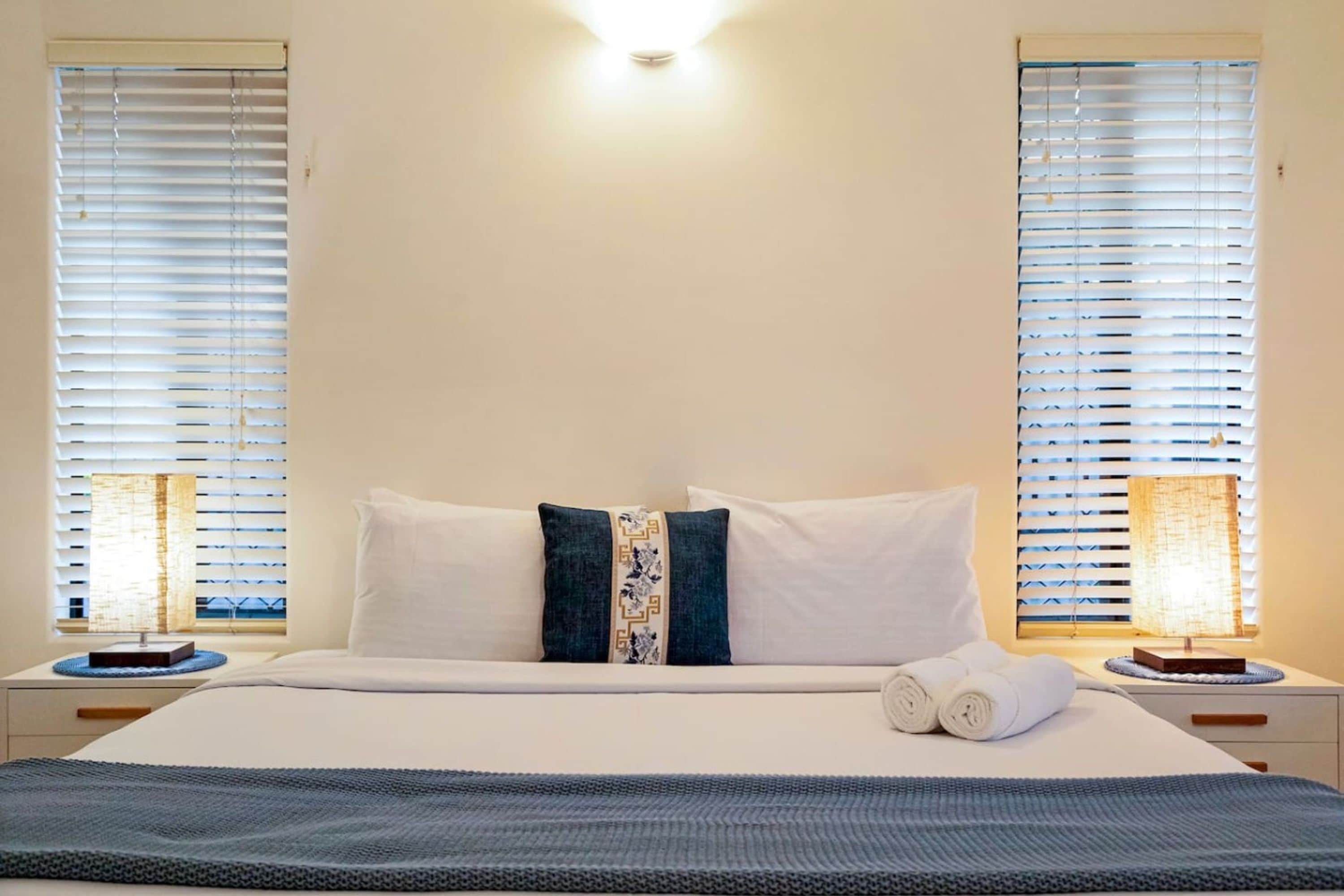 A hotel room with a bed, two bedside lamps, a window with blinds, and a wall-mounted light fixture.