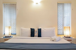 A hotel room with a bed, two bedside lamps, a window with blinds, and a wall-mounted light fixture.