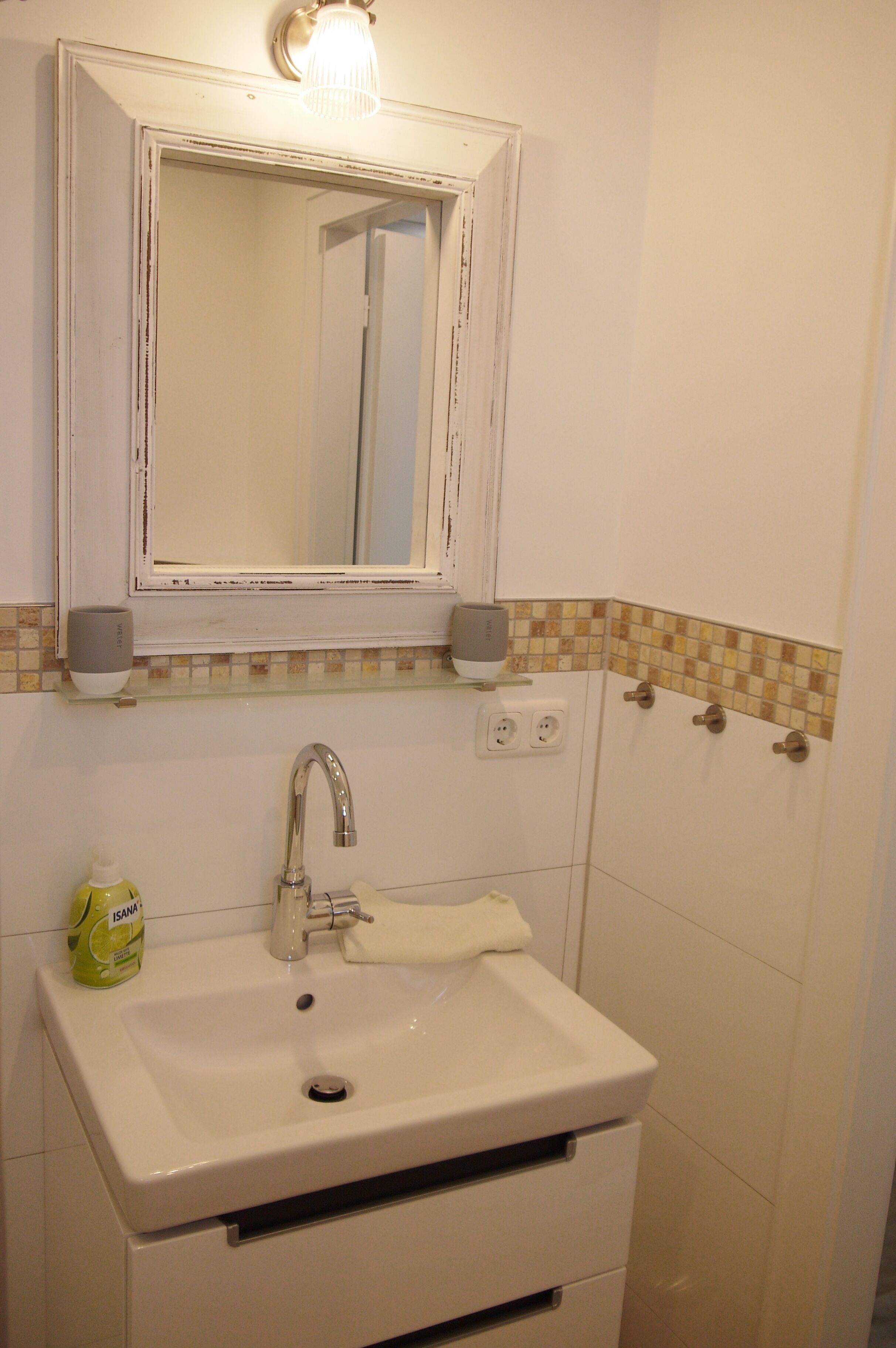 Un baño con un lavamanos blanco, un espejo encima y una lámpara. Hay dos ganchos para toallas en la pared y un dispensador de jabón.