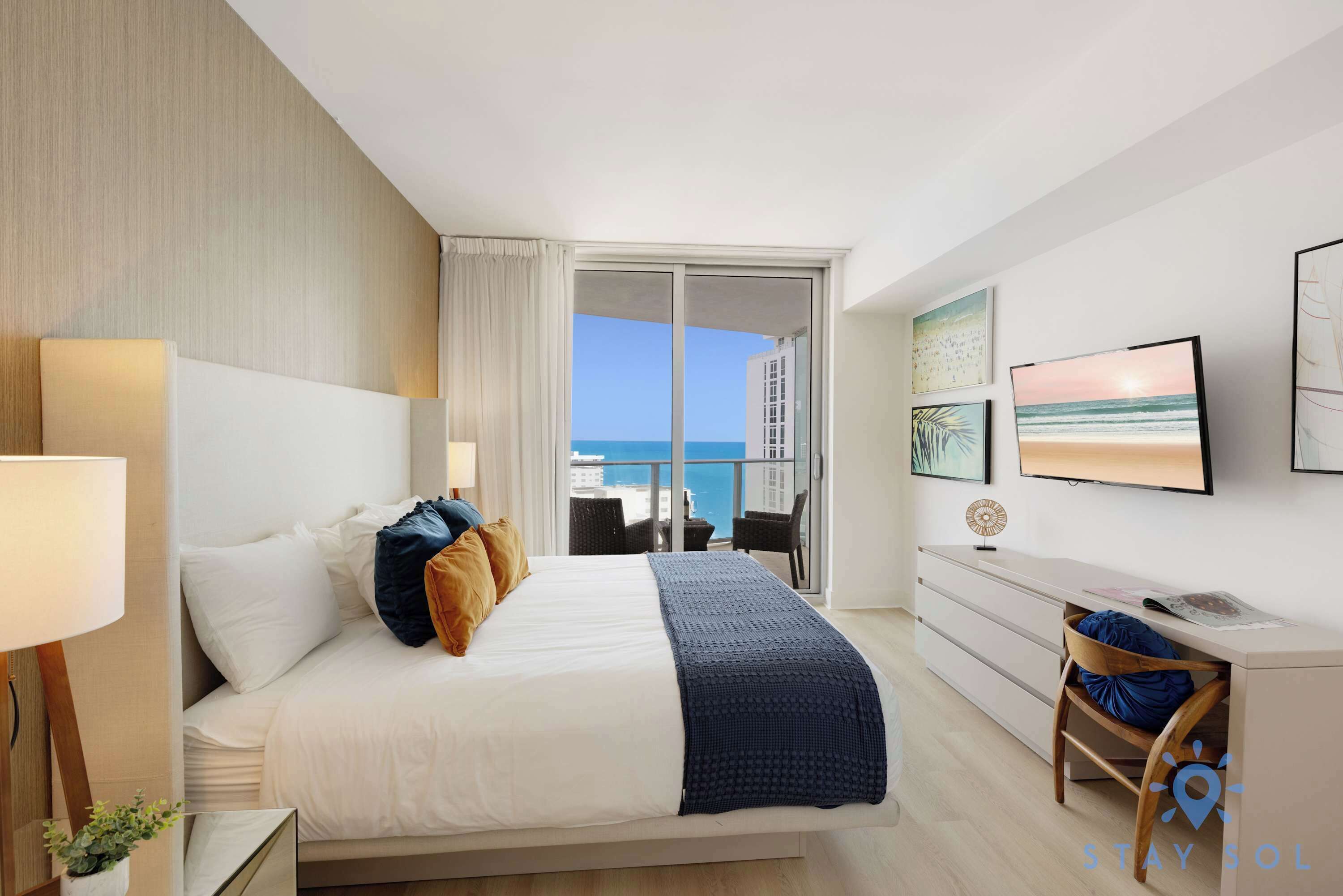 A hotel room with a bed, a desk, a chair, a lamp, and a view of the ocean.