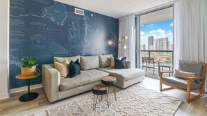 Ein modernes Wohnzimmer mit einem Ecksofa, einem kleinen runden Tisch und einem Sessel. Ein großes Fenster bietet Blick auf die Stadt.