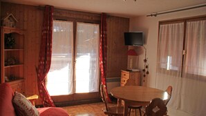 A room with a wooden floor, a round wooden table with chairs, a television mounted on the wall, and a sliding glass door with curtains. - Gelinote Chalet for 4 People (Chatel)