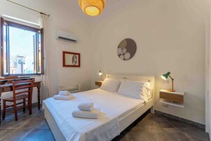 Habitación de hotel con cama, escritorio, silla, mesita de noche, aire acondicionado en la pared y una ventana con cortinas.
