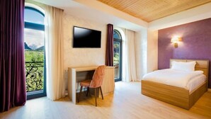 Une chambre à coucher avec un lit, un bureau et un téléviseur. Il y a une fenêtre avec des rideaux et une vue sur la verdure.