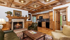 Un salon chaleureux avec une cheminée, un plafond en bois et une cuisine à l’arrière-plan.
