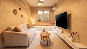 A modern living room with a sofa, a small wooden table, a flat-screen TV, and a window with a view of greenery. - Gaia Nest 1-bedroom apartment in Piraeus (Pireas)